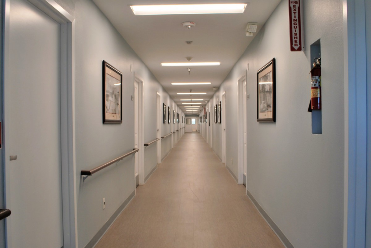 A long, well-lit corridor in a hospital or clinic with doors and framed pictures on the walls.