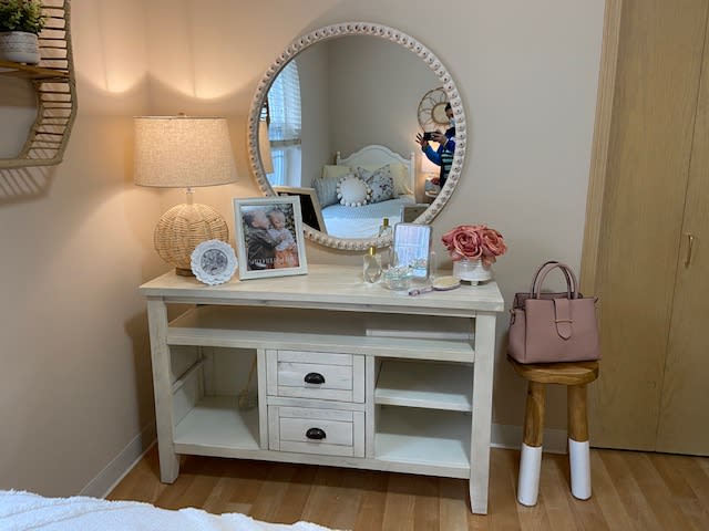 A well-decorated vanity with a round mirror and a table lamp, adorned with flowers, photo frames, and a handbag.