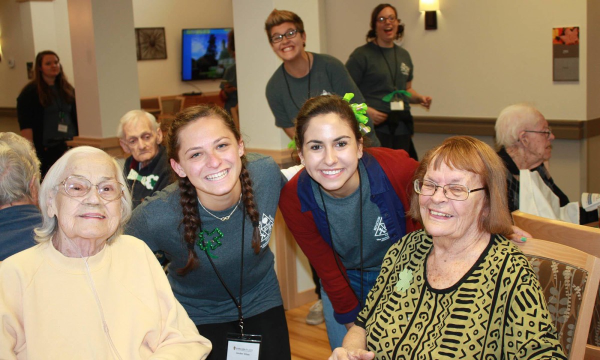 A group of smiling people, including seniors and volunteers, gathered indoors in a community setting.