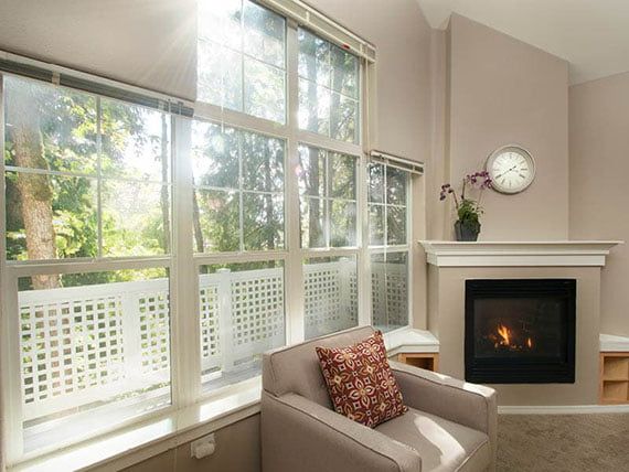A cozy living room with a fireplace, large windows, and an armchair bathed in daylight.