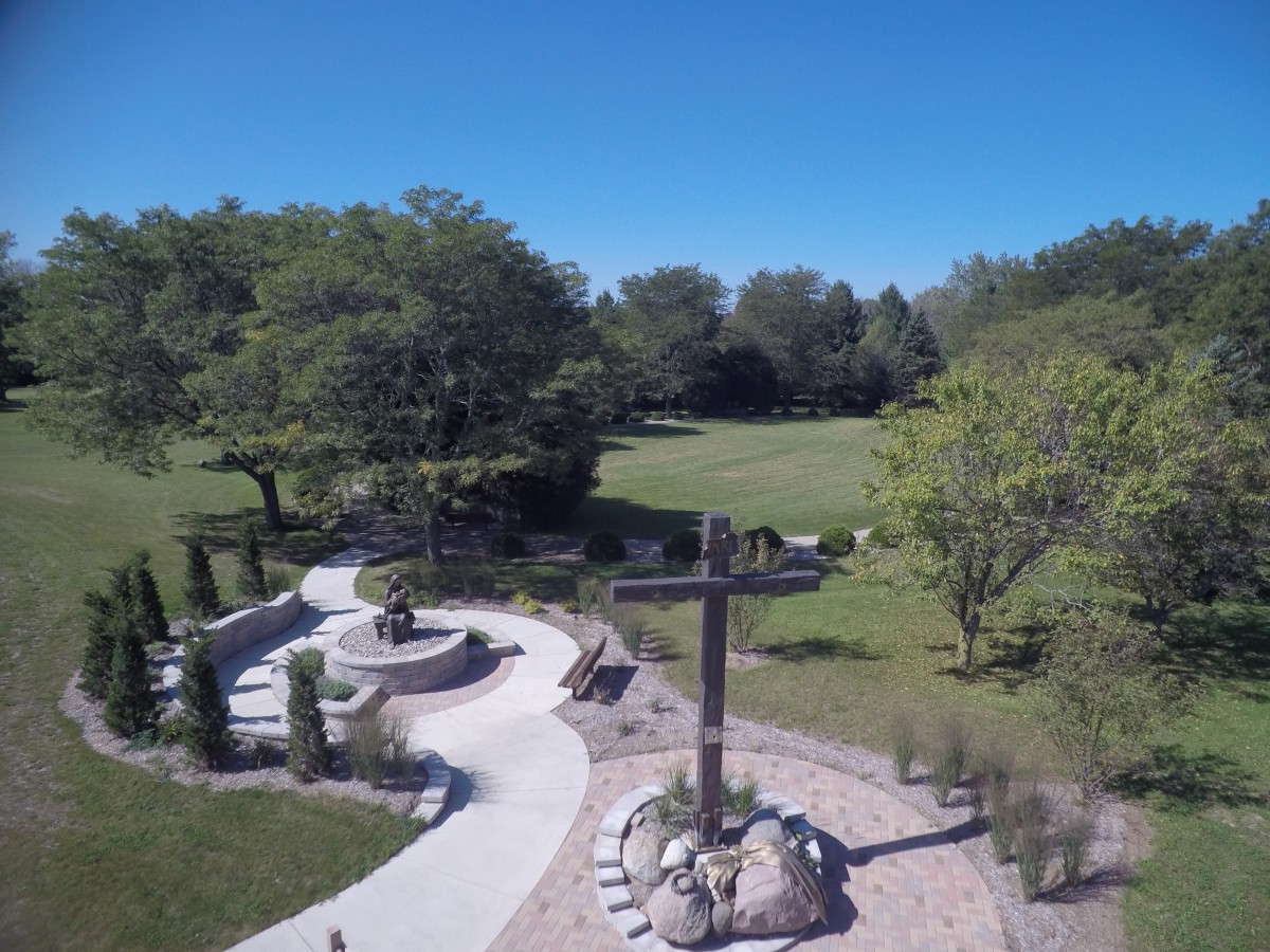 A serene outdoor pathway featuring a large cross, surrounded by trees and greenery.