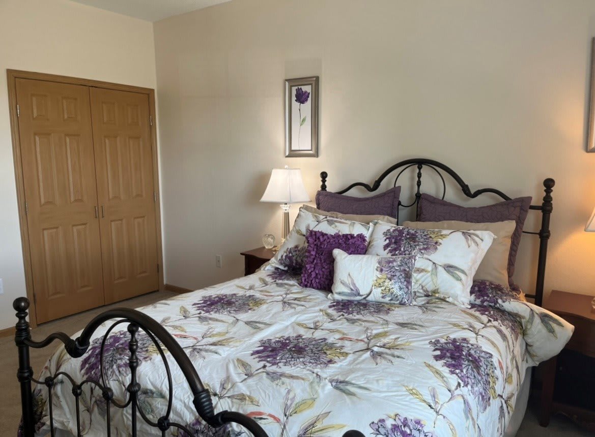 A cozy bedroom with floral bedding and an iron bed frame, featuring a nightstand with a lamp and a closet in the corner.