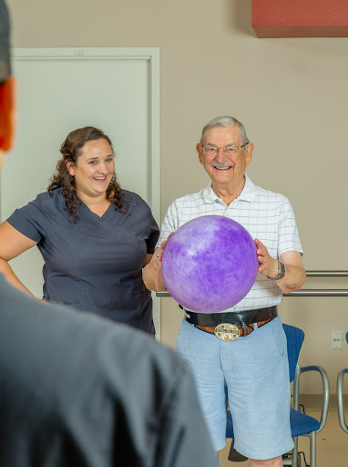 An elderly man joyfully holds a purple ball while smiling at someone off-camera, alongside a woman who is also smiling.