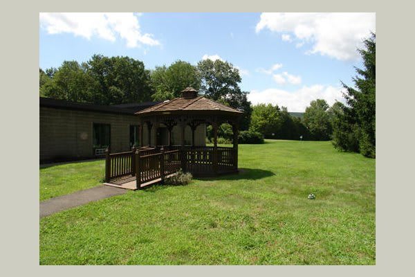 A scenic garden with a wooden gazebo surrounded by a lush green lawn and trees under a partly cloudy sky.