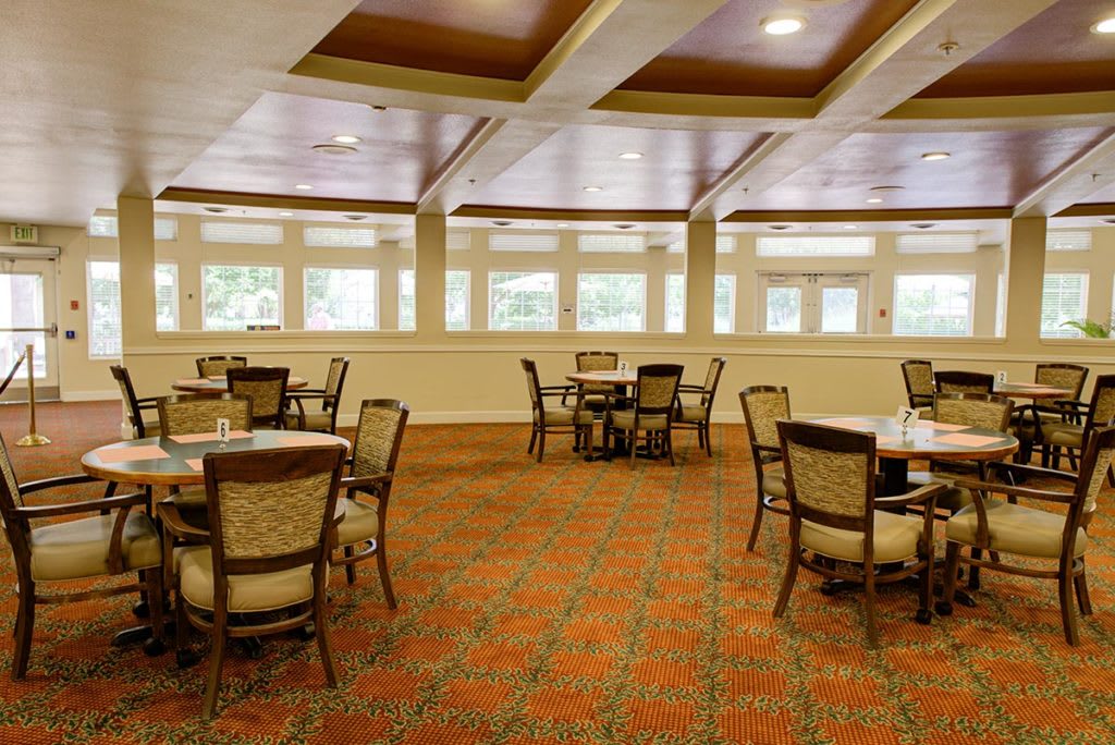 A spacious dining area with multiple tables and chairs, featuring large windows and patterned carpet.