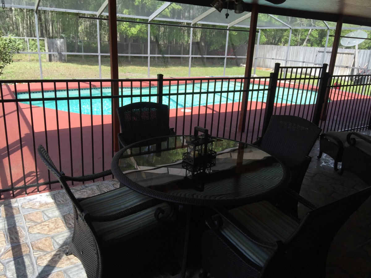 A peaceful backyard scene featuring a swimming pool, bordered by a fence, with patio furniture set under a covered area.