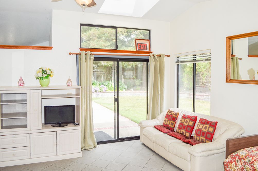 A modern living room with a white sofa adorned with red pillows, a small television on a stand, and large sliding glass doors leading to an outdoor view.