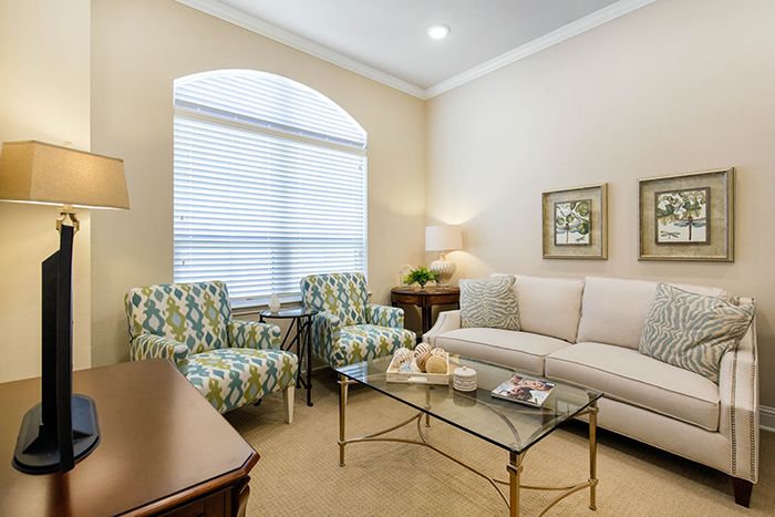 A cozy living room featuring a sofa, patterned armchairs, and a glass coffee table with decorative items. A large window allows natural light to illuminate the space.