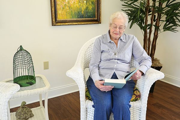 An elderly woman sitting in a chair reading a book indoors.