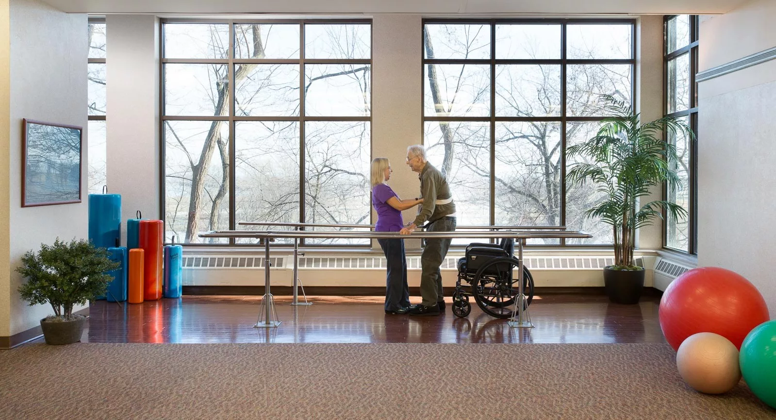 An elderly person receiving physical therapy assistance in a spacious room with large windows.