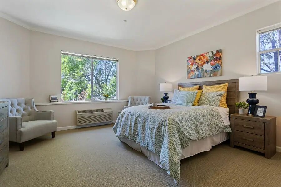 A cozy bedroom with soft lighting, featuring a neatly made bed, artwork on the wall, and a large window letting in natural light.