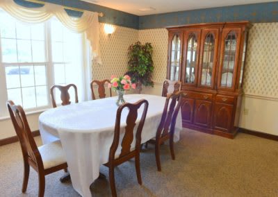 A traditional dining room with a table covered by a white cloth, surrounded by wooden chairs and featuring a glass-fronted cabinet.