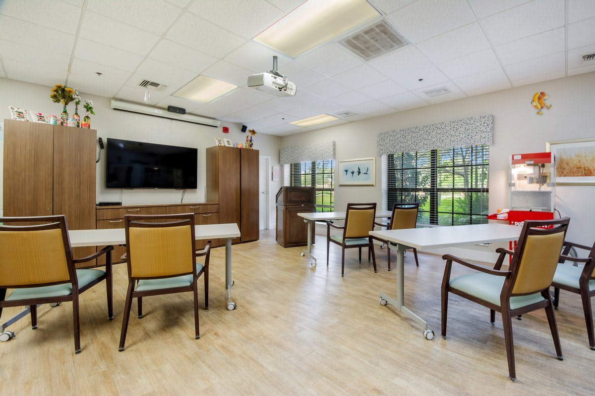 A well-lit classroom with tables, chairs, a TV screen, and large windows.
