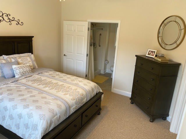 A cozy bedroom featuring a neatly made bed with patterned bedding, a wooden dresser, and an en suite bathroom doorway.