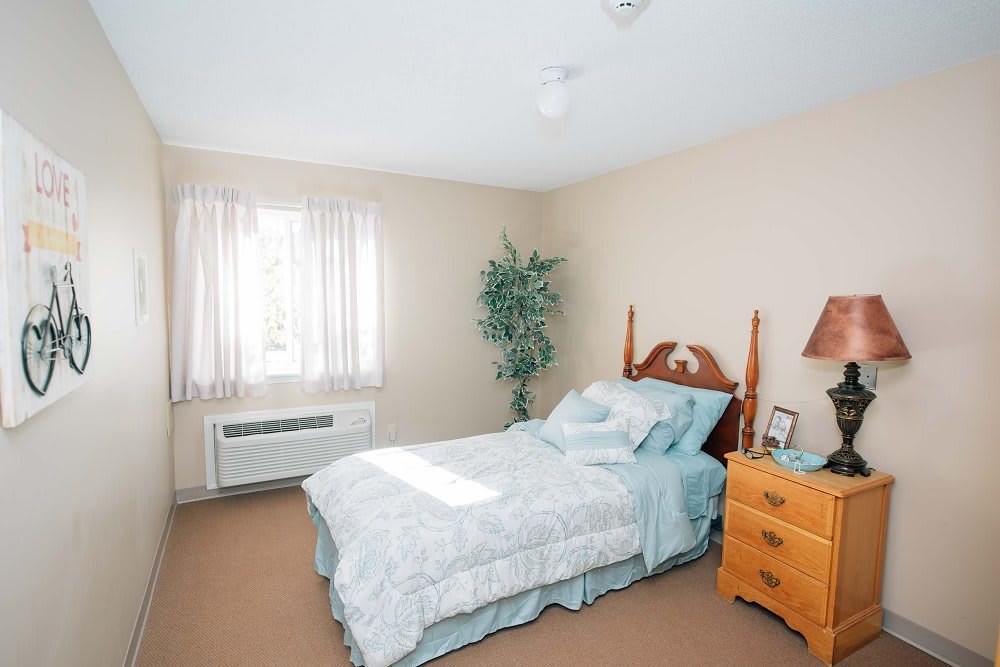 A cozy bedroom featuring a single bed with light blue bedding, a wooden nightstand, and a large window with sheer curtains.