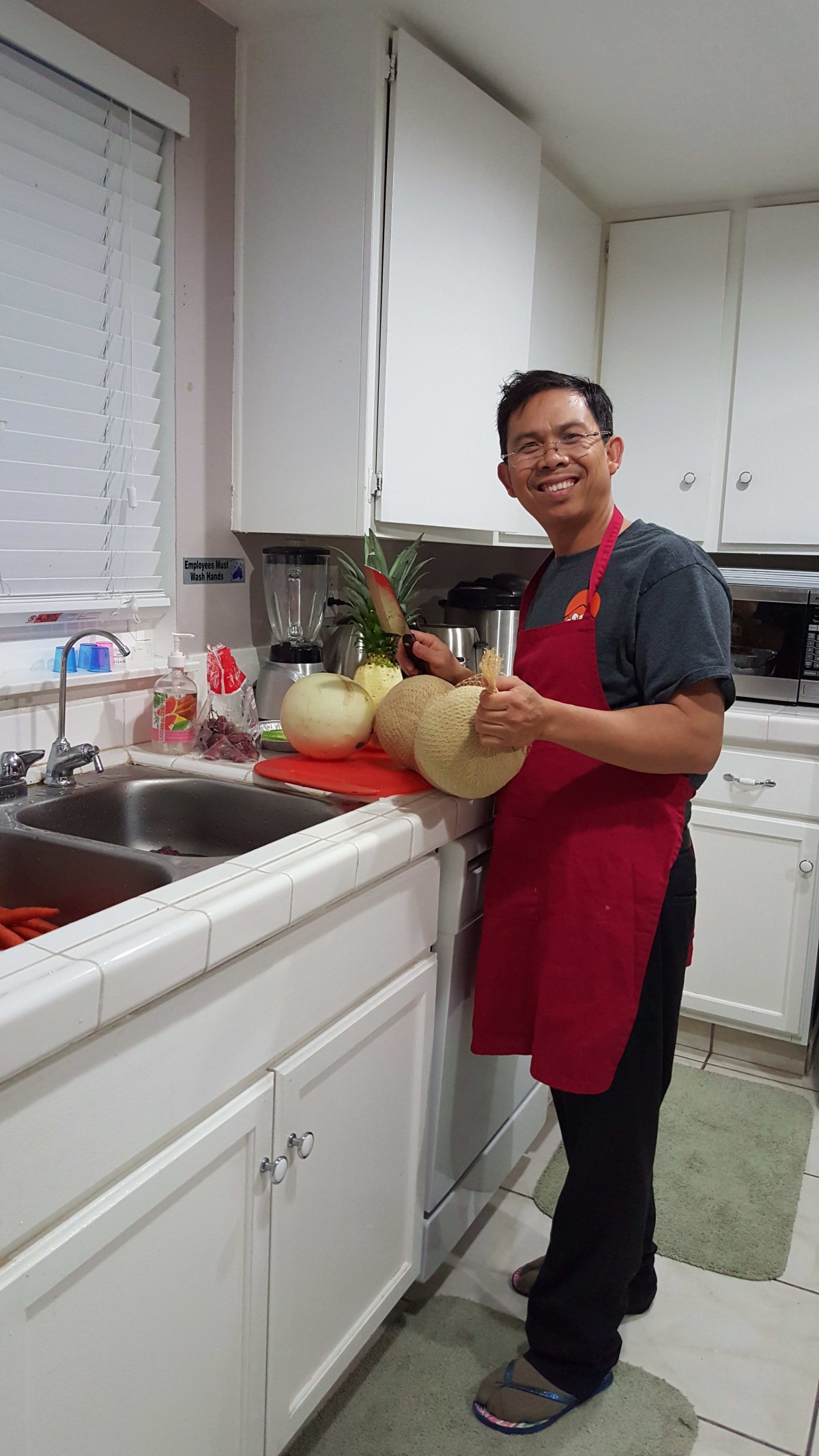 A person in a kitchen smiling while preparing melons on a cutting board.