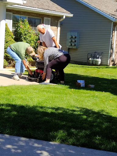 A group of people collaborating on a lawn mower repair in a sunny yard.