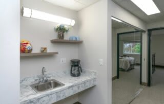 A small kitchenette with a sink, coffee maker, and shelves in a bright room.