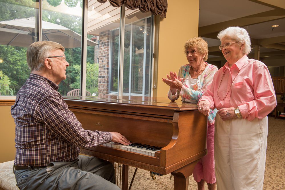 A group of elderly friends enjoying music, with one person playing the piano while others smile and clap indoors.