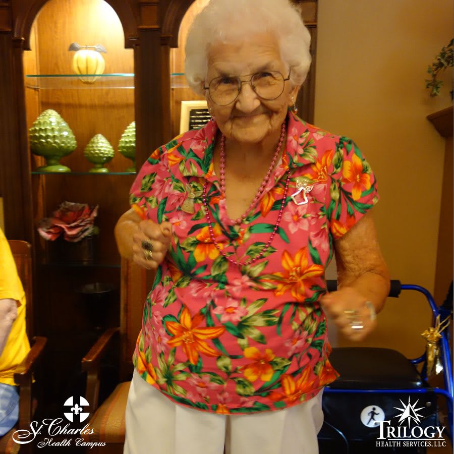 An elderly woman wearing a vibrant floral shirt and glasses is smiling indoors, with a walker nearby.