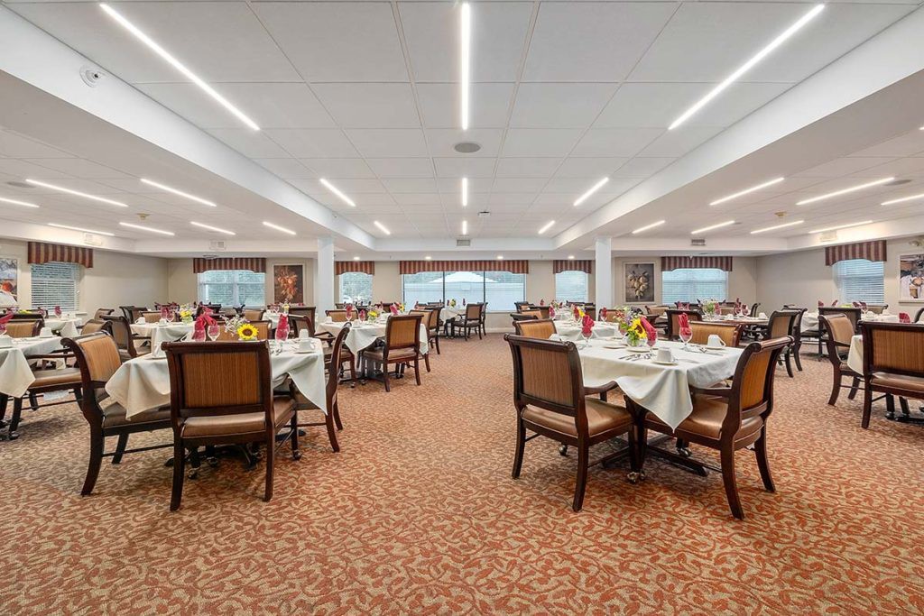 A spacious dining room with neatly arranged tables and chairs, featuring warm lighting and a carpeted floor.