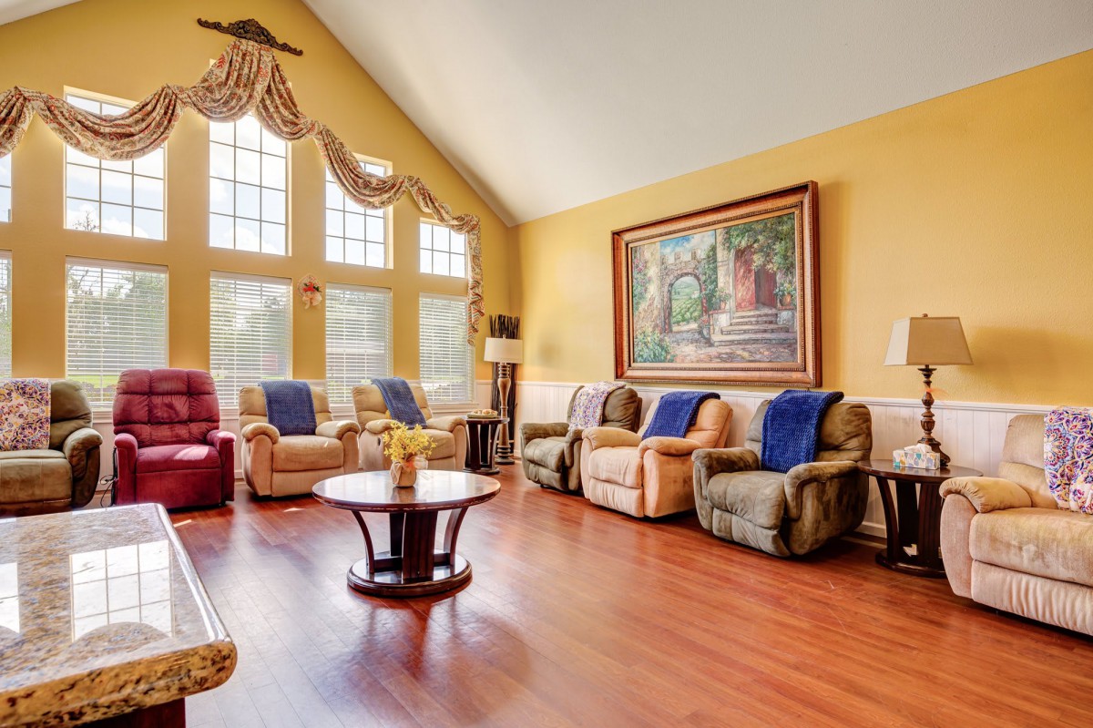 A spacious living room with warm yellow walls, comfortable armchairs, and a large wall painting under abundant natural light from tall windows.