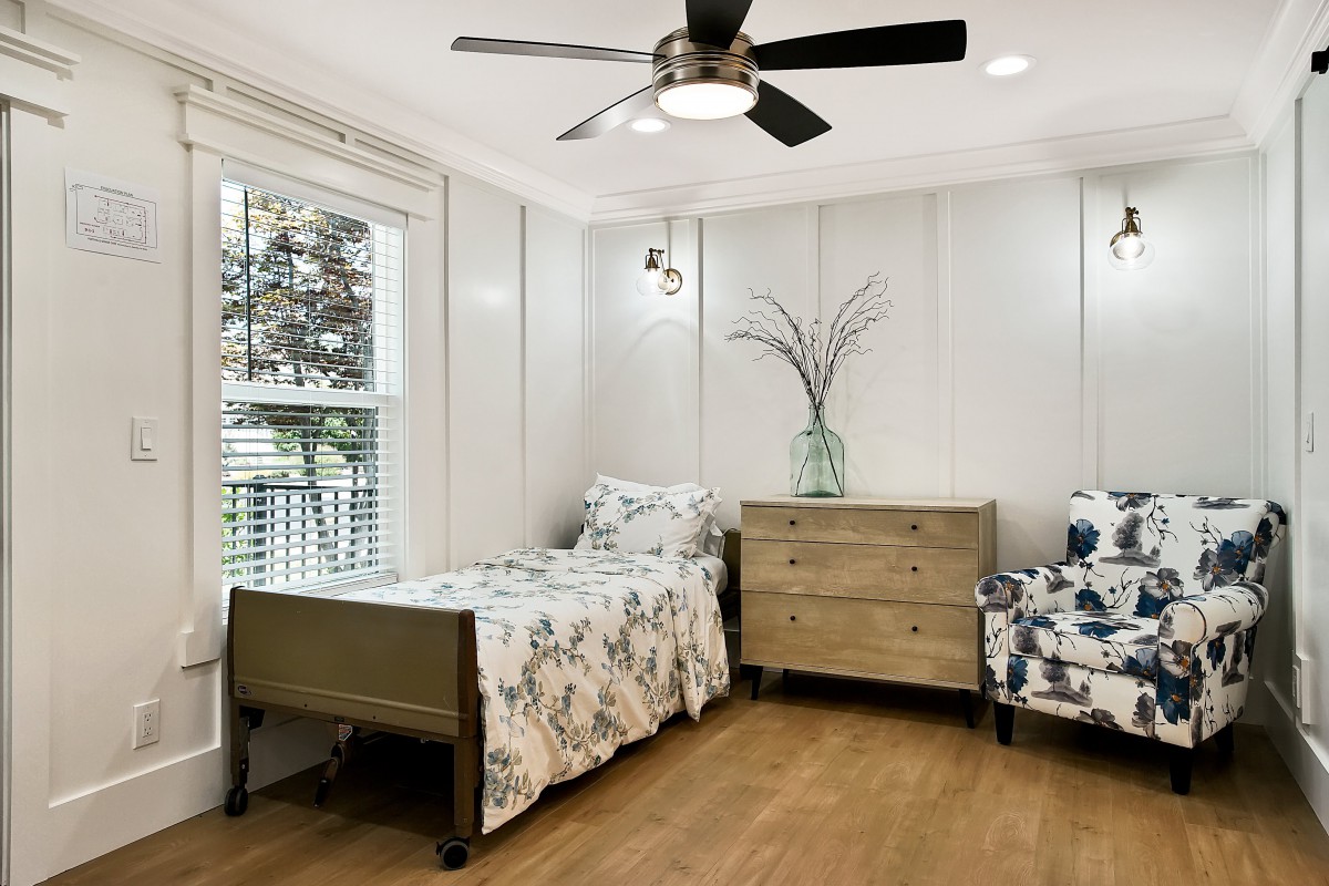A cozy bedroom with a single bed, wooden dresser, and floral armchair, featuring a ceiling fan and large window.