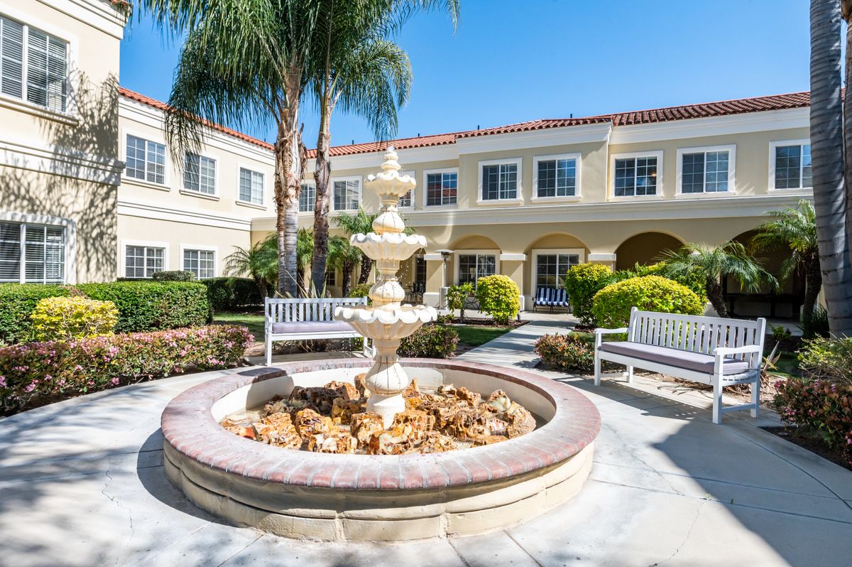 A sunny courtyard featuring a central fountain, surrounded by benches and greenery, with a building in the background.