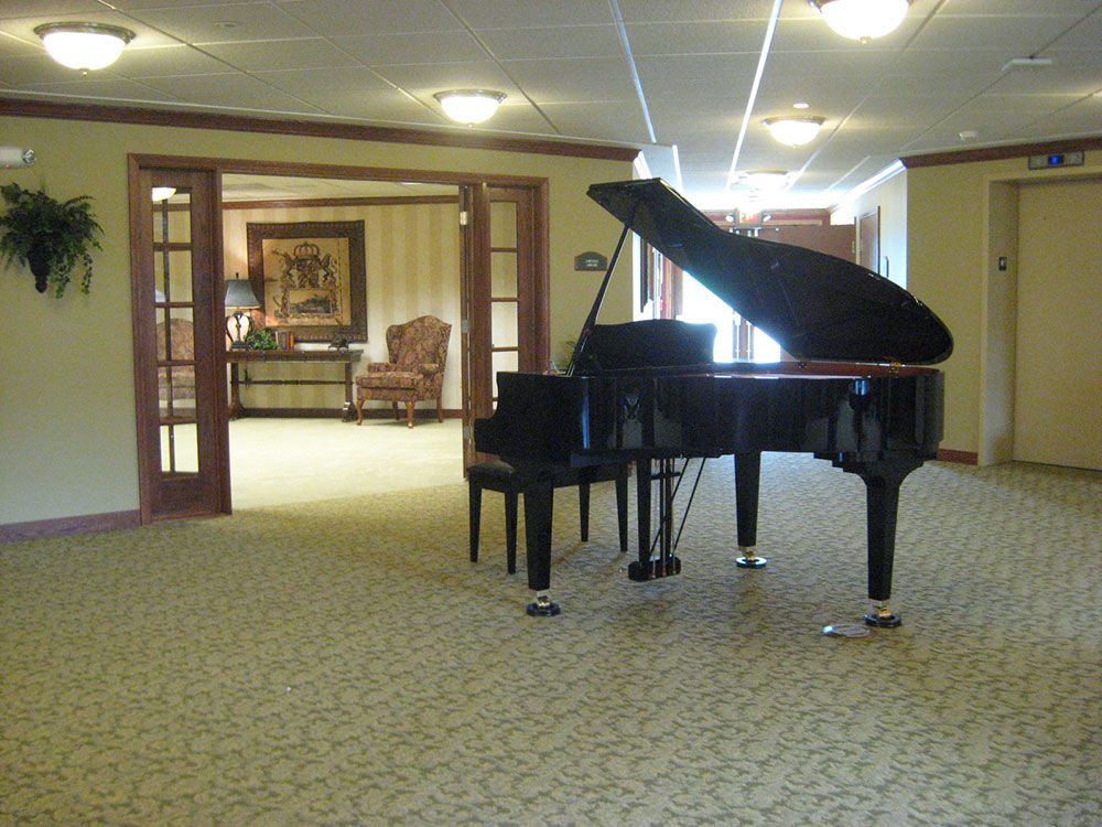 A grand piano is situated in a carpeted living room with an open doorway leading to a cozy seating area.
