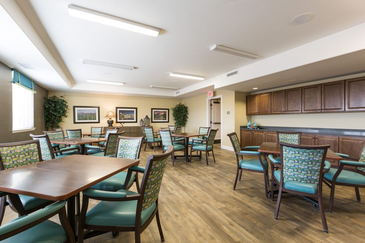 A spacious dining area with wooden floors, multiple tables and chairs, decorated with paintings and plants.