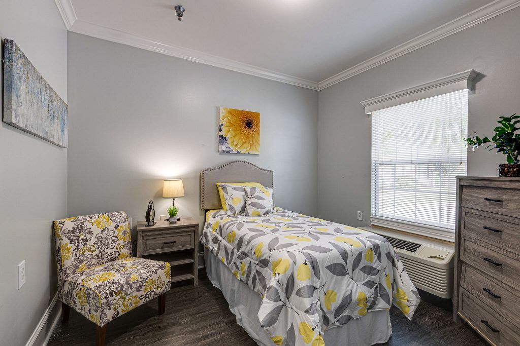 A cozy bedroom with a single bed featuring floral bedding, complemented by an armchair and a nightstand with a lamp.