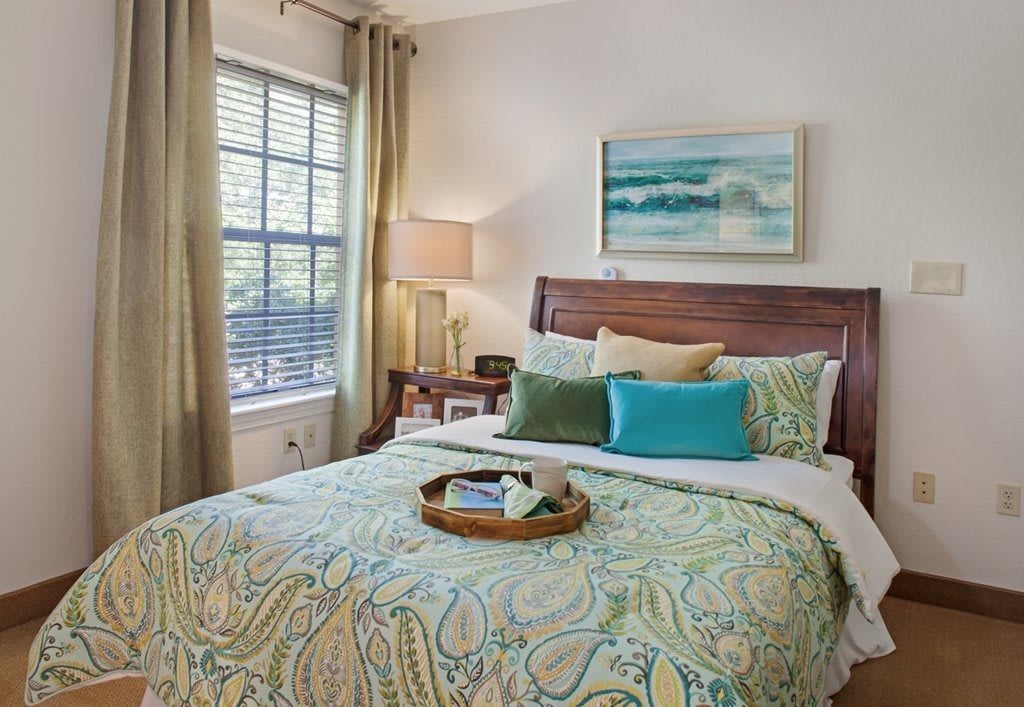 A cozy and well-decorated bedroom featuring a bed with patterned bedding, colorful pillows, a bedside table with a lamp, and a window with curtains. A painting hangs above the bed.