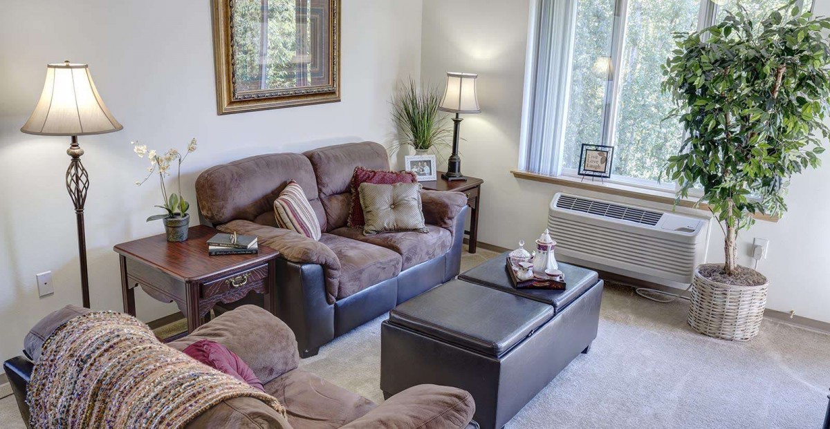 A cozy living room with a sofa, armchair, coffee table, and a lamp by the window.