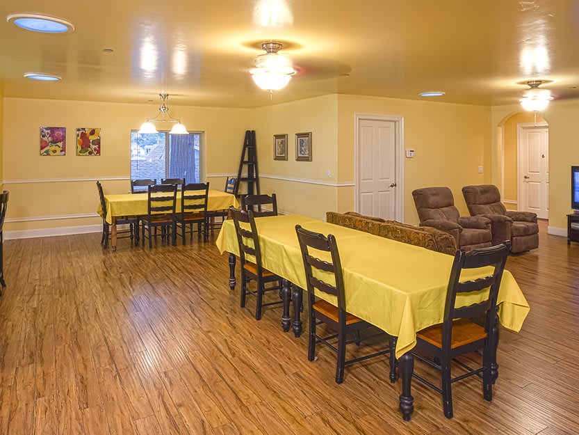 A dining room with tables covered in yellow tablecloths and wooden chairs on a wood floor.