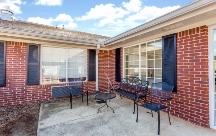 An outdoor patio area with metal chairs and a table, set against a brick wall with large windows.