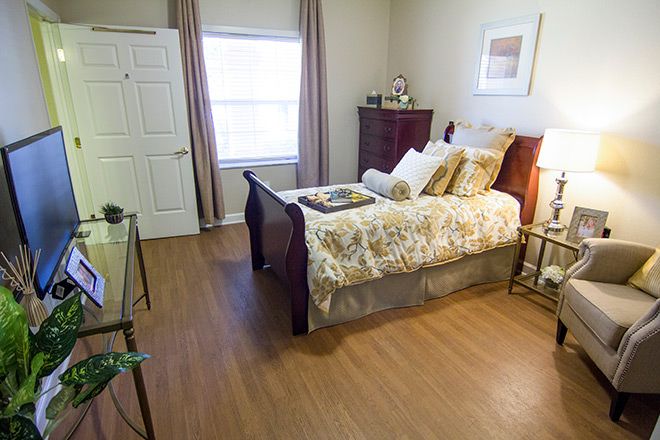 A cozy and well-decorated bedroom with wooden flooring, featuring a neatly made bed, bedside lamp, chest of drawers, and a window with curtains.