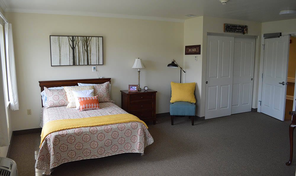 A cozy bedroom featuring a neatly made bed with colorful pillows, a bedside table with a lamp, and a comfortable chair in the corner.