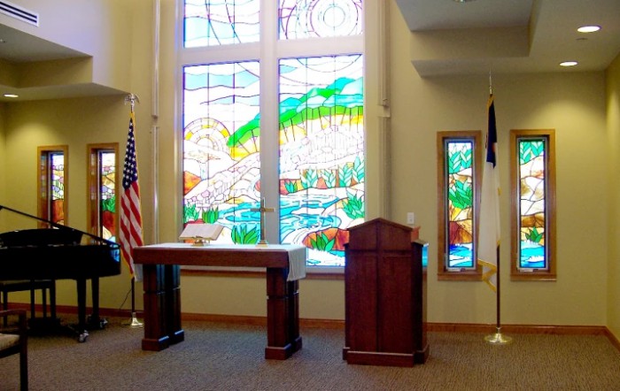 A chapel interior featuring stained glass windows, a podium, an altar, and flags.