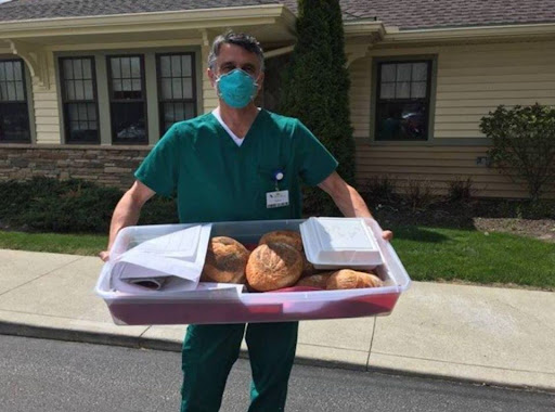 A medical professional wearing scrubs and a face mask holds a tray of freshly baked bread outside a healthcare facility.