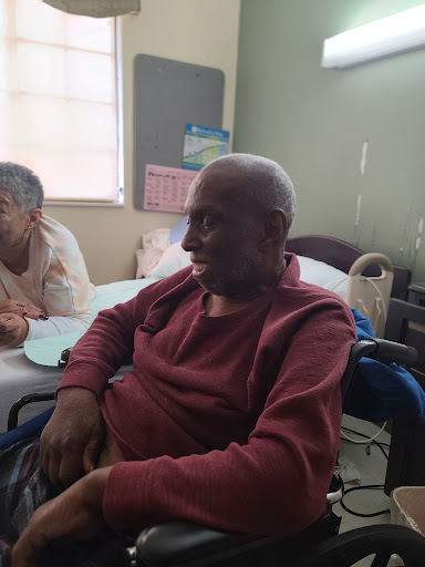 An elderly man sitting in a hospital room, smiling while another person is seen nearby.