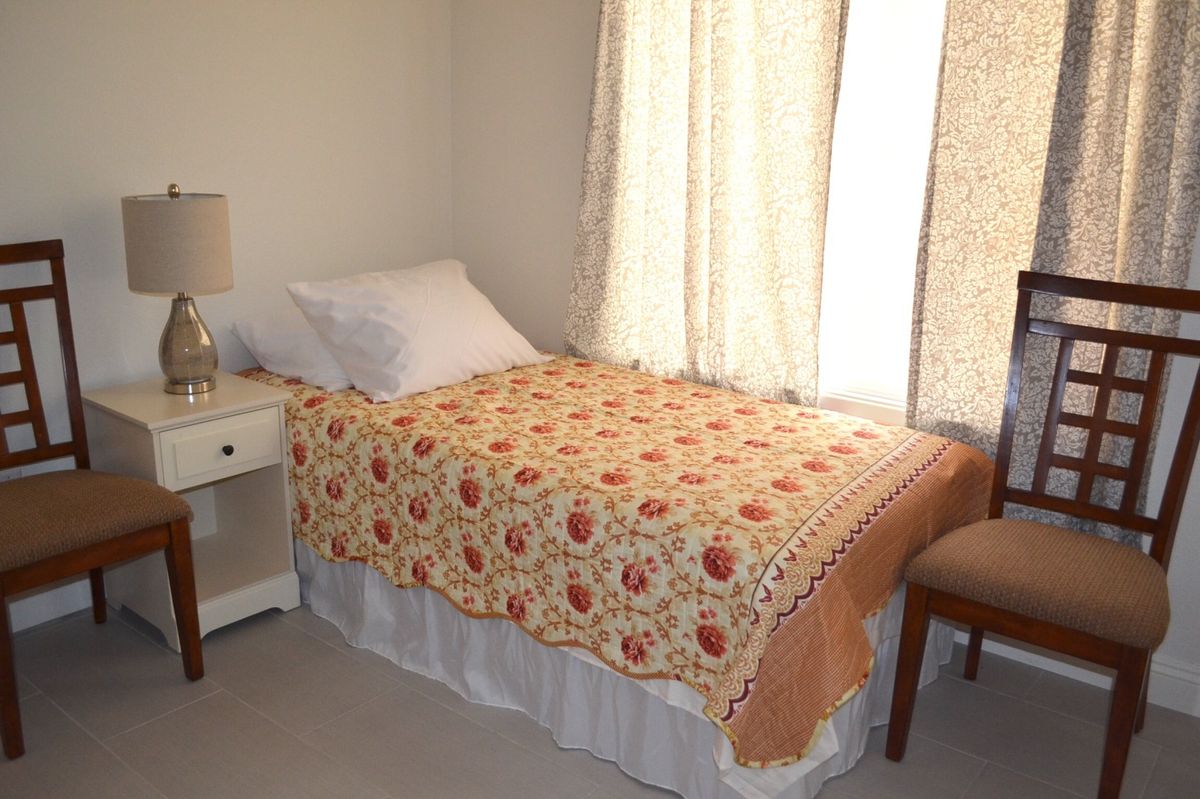A small bedroom with a single bed covered by a patterned quilt, next to a lamp and chair.