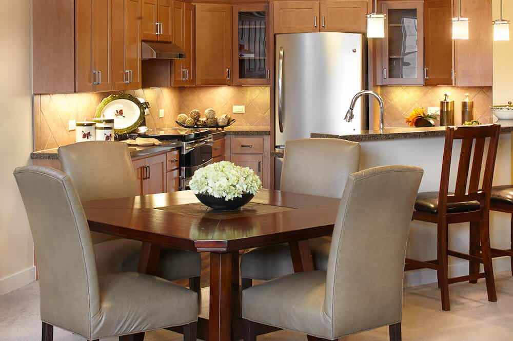A modern kitchen and dining area with wooden cabinets, a stainless steel refrigerator, and a central table with upholstered chairs.