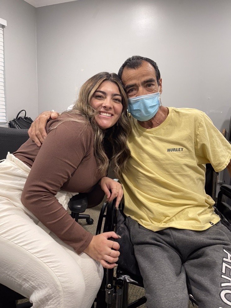 Two people smiling and hugging indoors, one wearing a mask.