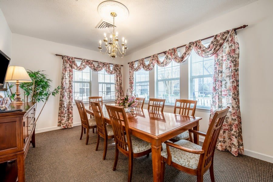 A well-lit dining room with a large wooden table, surrounded by elegant chairs and adorned with decorative curtains and a chandelier.