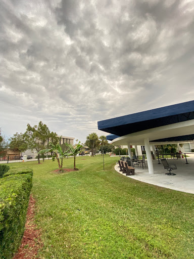 A scenic view of a park featuring lush green grass, trees, and a covered outdoor seating area under a cloudy sky.