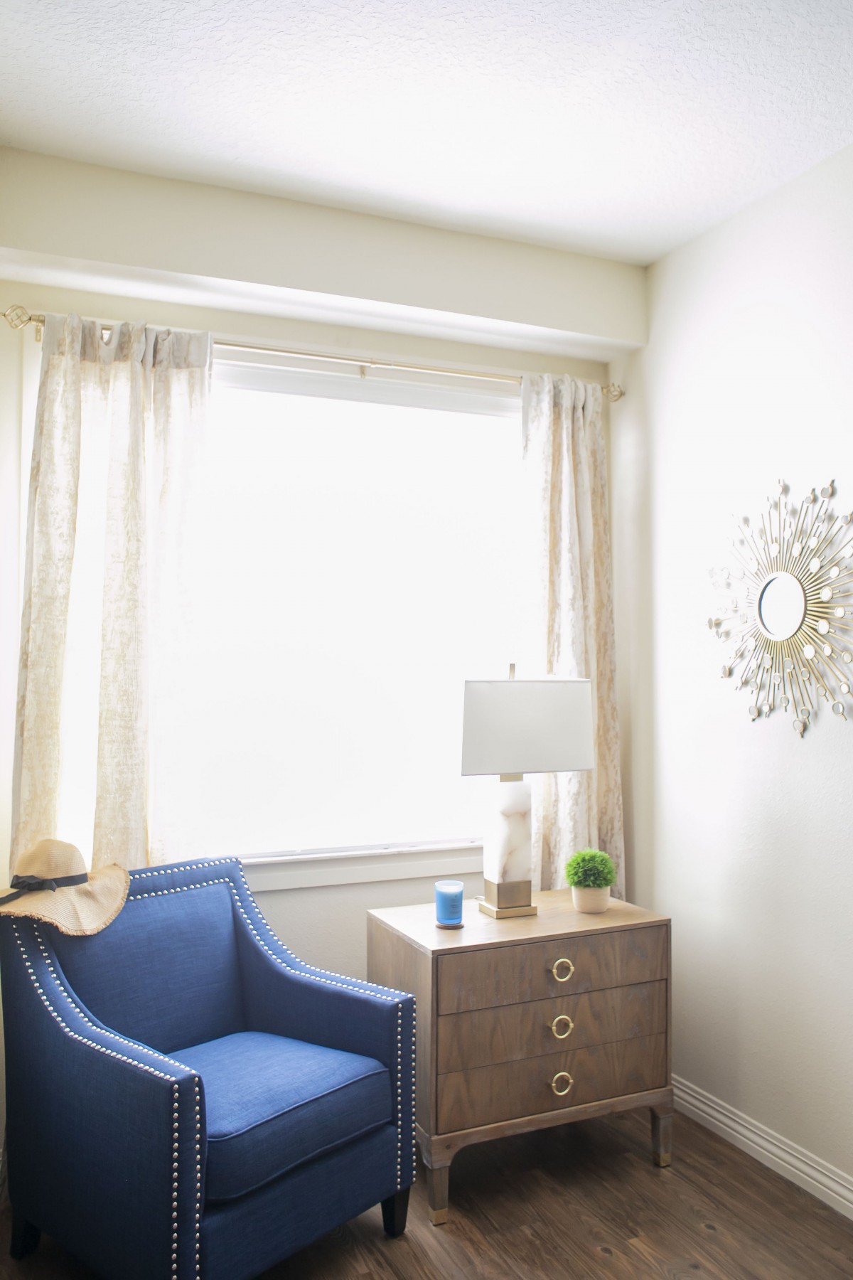 A cozy corner with a blue armchair, a side table with a lamp, and light curtains framing a large window.