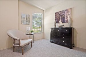 A cozy living room corner with a cushioned chair, a black dresser adorned with nautical decor, and a window letting in natural light.