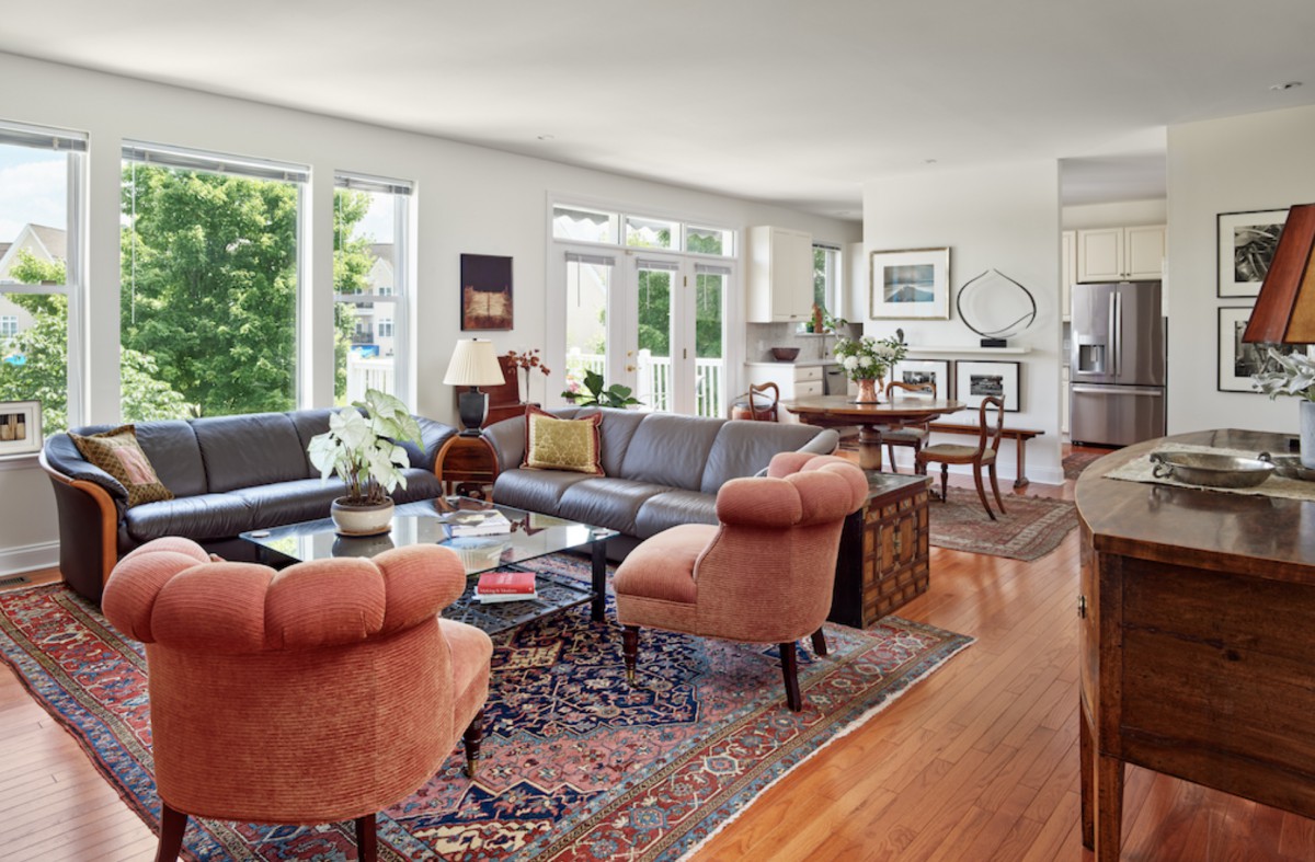 A spacious and well-lit living room featuring comfortable seating, a patterned rug, and large windows overlooking greenery.
