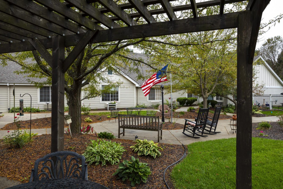 A serene garden patio with a bench and rocking chairs beneath a pergola, adorned with an American flag.
