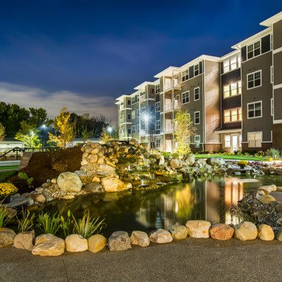 A modern apartment complex with a illuminated pond and landscaped surroundings at night.